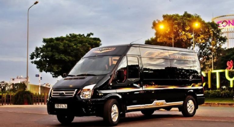 Xe khách Green Limo hiện đại, màu đen xanh, gần cây thông Noel và tòa nhà cao tầng.