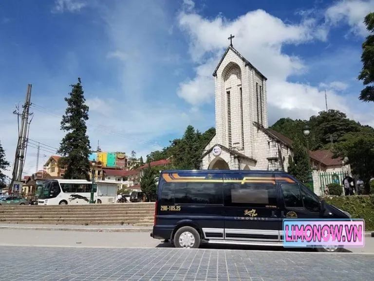 Xe limousine đen chất lượng cao đỗ trước Nhà thờ Đá Sapa, cảnh quan thị trấn núi phía sau.